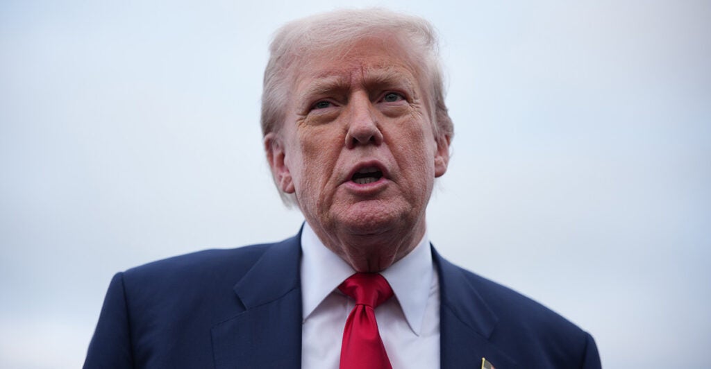 Close-up of President Donald Trump in blue suit and red tie against gray sky.