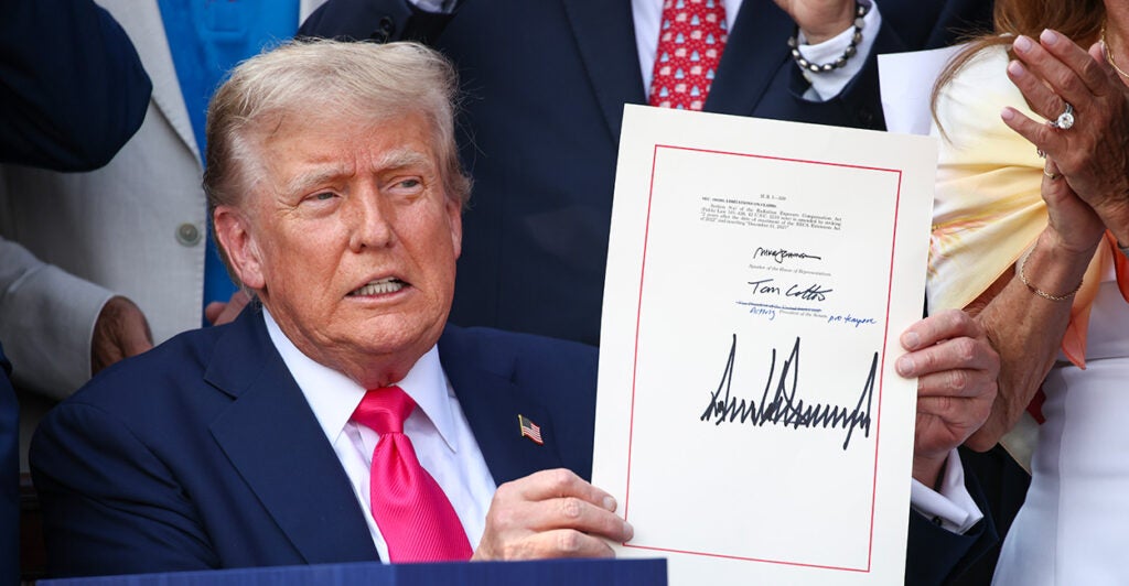 President Trump sitting at a desk holds up signed One Big Beautiful Bill Act.