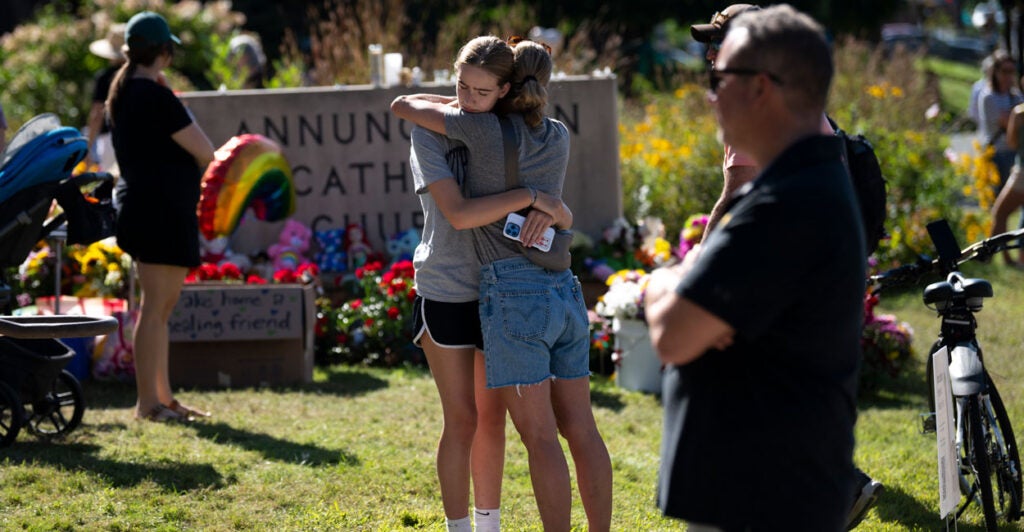 Two teen girls hug in front of a memorial of flowers at Annunciation Catholic Church.
