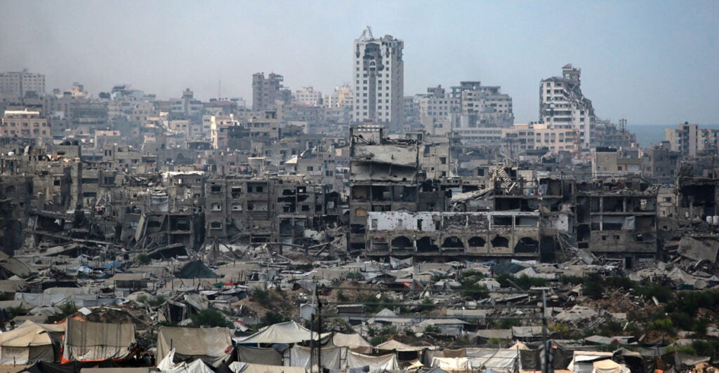 The war torn skyline of Gaza City is shown with tents set up beneath crumbling buildings.