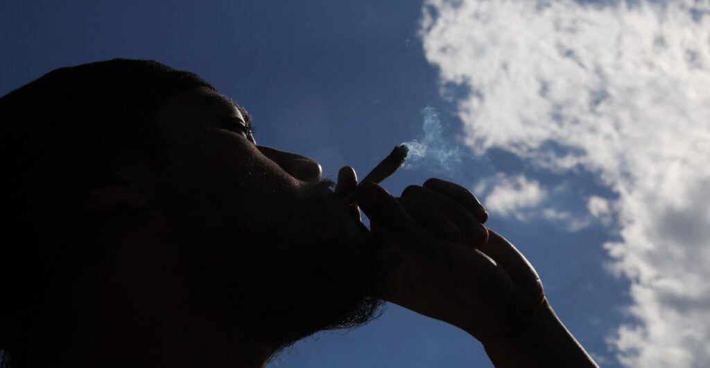 A pot smoker is silhouetted against a blue sky, with the only cloud being a cloud of marijuana smoke