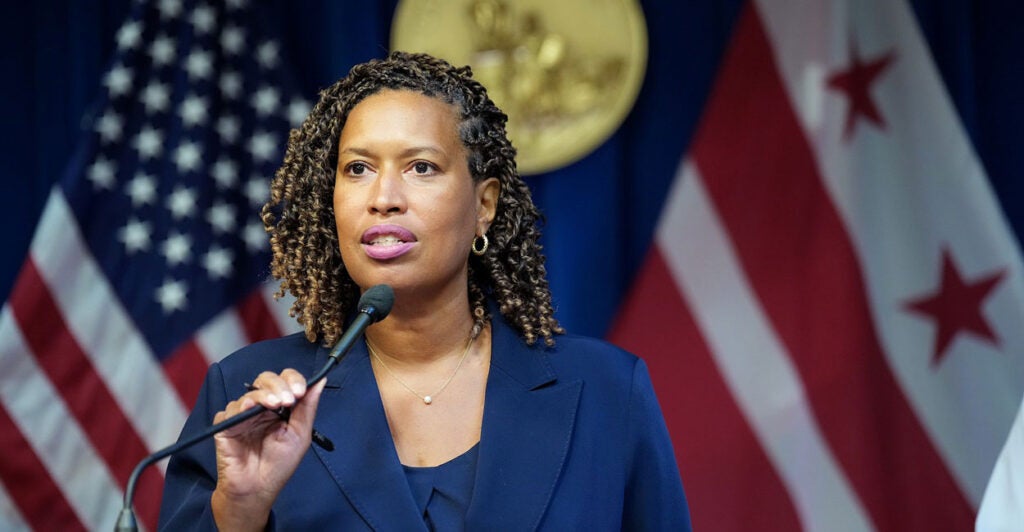Muriel Bowser stands in front of a podium and holds the microphone with her right hand as she begins to talk.
