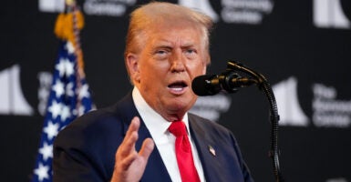 Donald Trump stands before a microphone while talking and gestures with his right hand.
