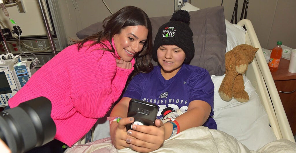Lea Michele leans next to a boy wearing a knit cap in a hospital bed for a selfie.
