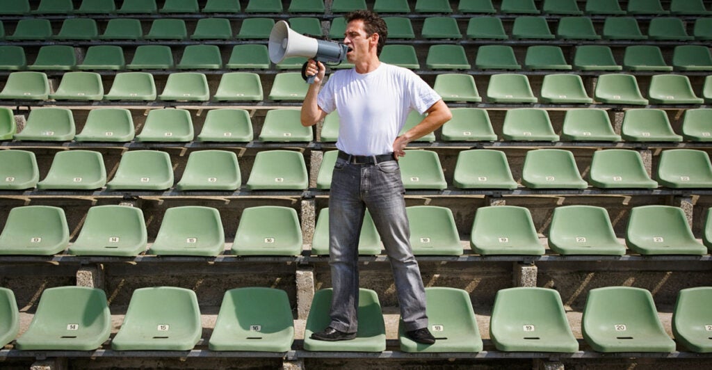 A man in a white shirt and jeans stands on seats surrounded by empty seats, shouting into a megaphone.