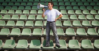 A man in a white shirt and jeans stands on seats surrounded by empty seats, shouting into a megaphone.