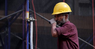 A bearded construction worker in yellow hard hat and burgundy shirt rests against a horizontal pole.