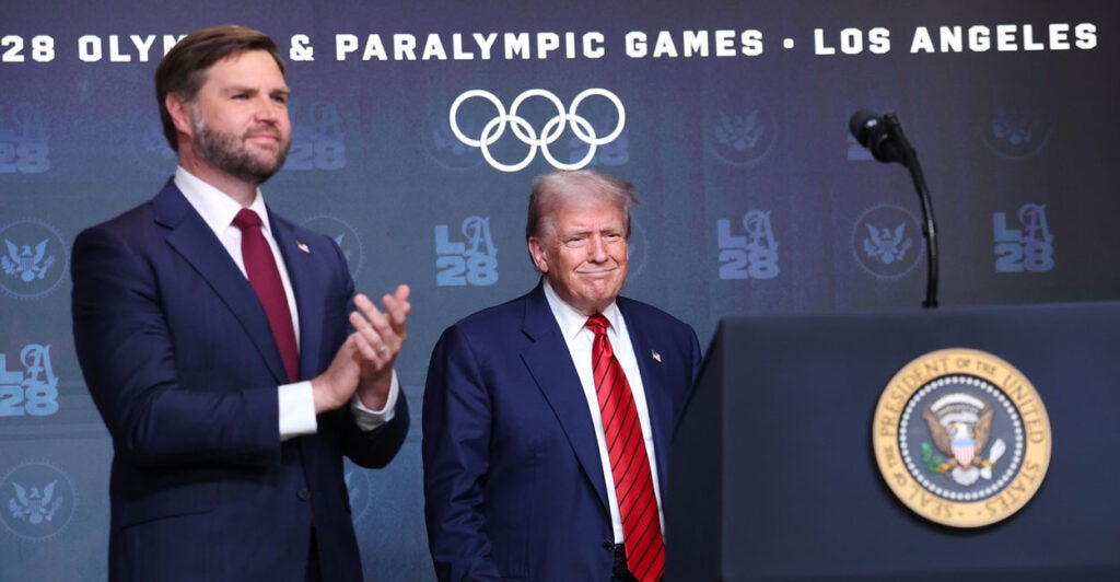 President Donald Trump and Vice President JD Vance arrive at a podium with the Olympic logo in the background.