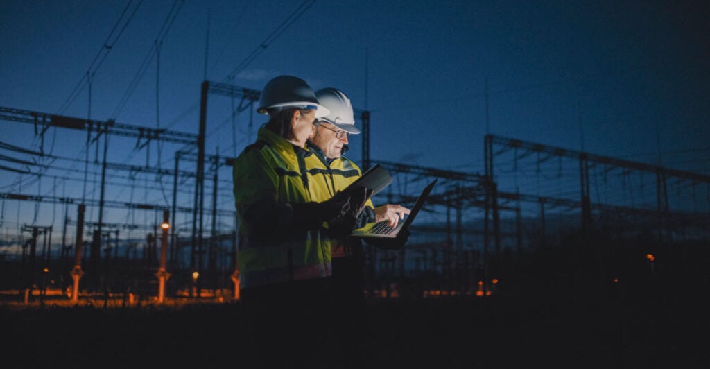 Two helmeted power plant workers talk as the plant is silhoutted against a darkening sky.