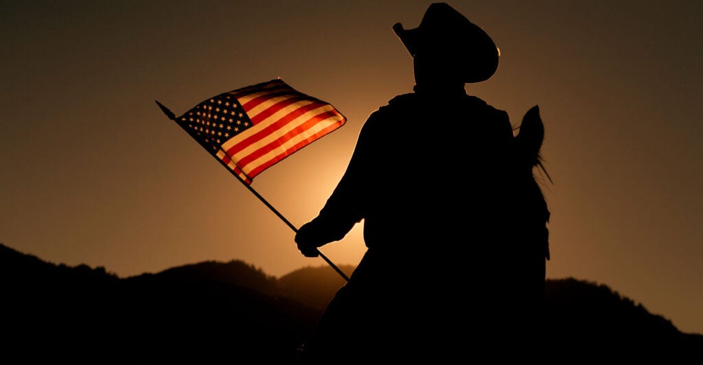 A cowboy on a horse in silhouette holds an American flag lit by the setting sun.
