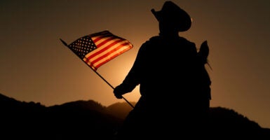A cowboy on a horse in silhouette holds an American flag lit by the setting sun.