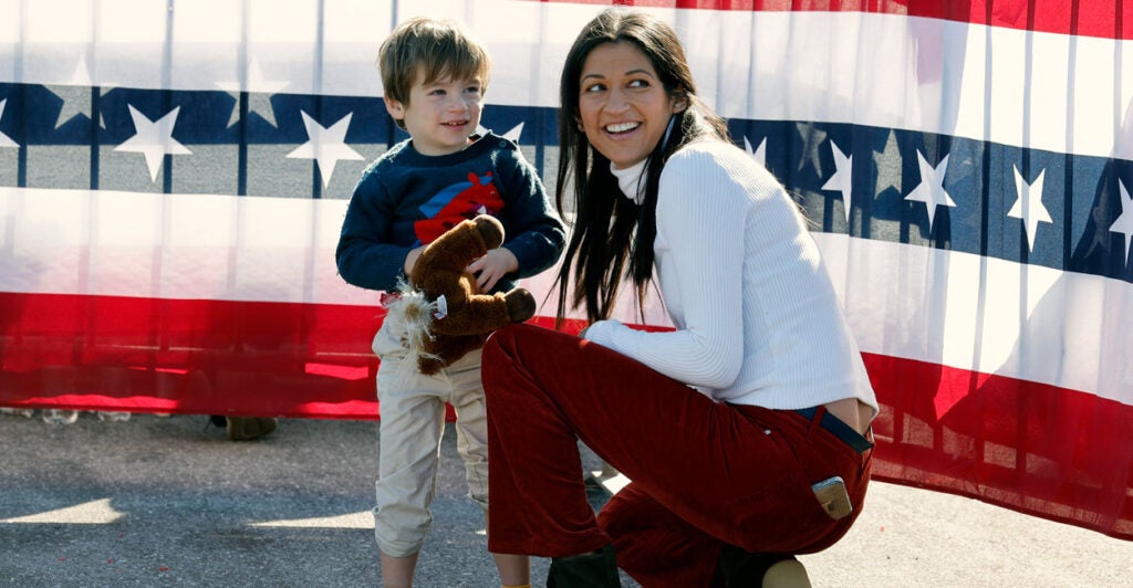 A smiling Katie Miller, in white turtleneck and burgundy pants, kneels with her young son in front of stars-and-stripes bunting.