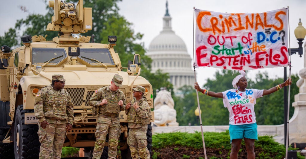Woman standing next to National Guard members and their vehicle holds sign reading "Criminals Out of DC? Start With the Epstein Files." The U.S. Capitol is in the background.