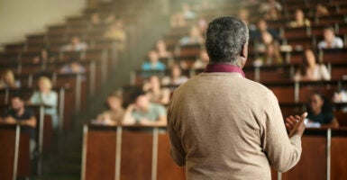 A black professor in tan sweater, with back to camera faces a lecture hall filled with students.