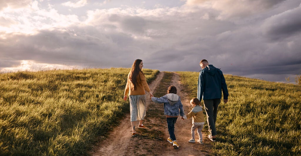 A mom and dad walk their two young children down a dirt road on an open plain, with a grand mostly cloudy sky ahead.