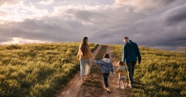 A mom and dad walk their two young children down a dirt road on an open plain, with a grand mostly cloudy sky ahead.