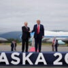 President Donald Trump and Russian President Vladimir Putin shake hands on a stage that says 'ALASKA 2025' on the front of it. Air Force One and fighter jets sit in the background.