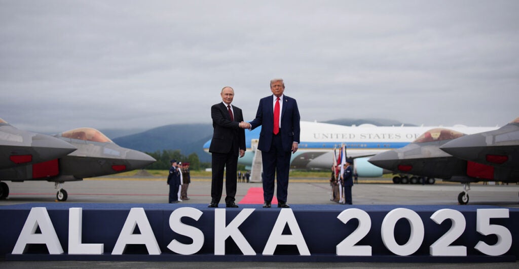 President Donald Trump and Russian President Vladimir Putin shake hands on a stage that says 'ALASKA 2025' on the front of it. Air Force One and fighter jets sit in the background.