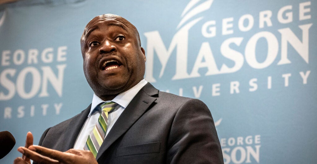George Mason University President Gregory Washington in a dark suit speaks at a press conference with the George Mason University logo in the background