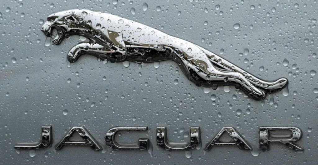 The photo features the logo for the luxury British car brand Jaguar up close, covered in rain drops. The logo has a leaping jaguar above the name "Jaguar."