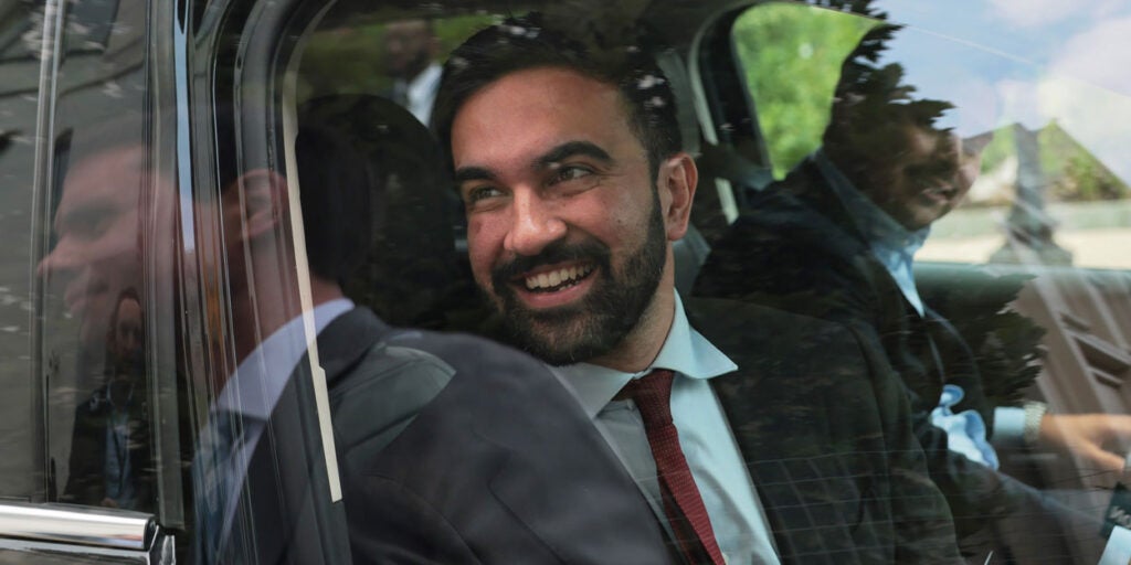 Zohran Mamdani smiles as he looks out the window of a car.
