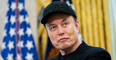 Elon Musk in a black T-shirt, black sport coat and black DOGE baseball cap in the oval office