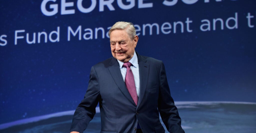 George Soros in a blue suit with purple tie against a blue background.
