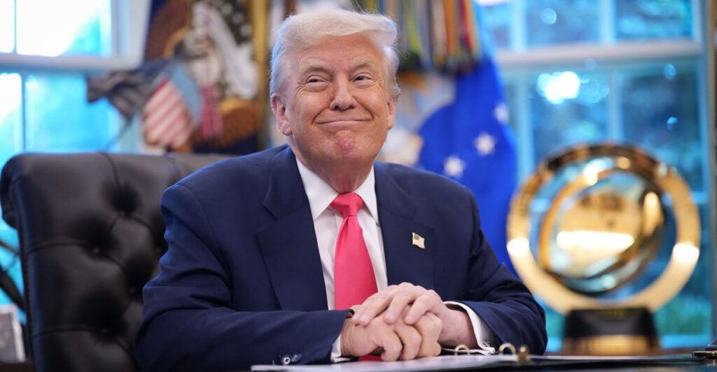 President Donald Trump smiles while sitting at his desk in the Oval Office.
