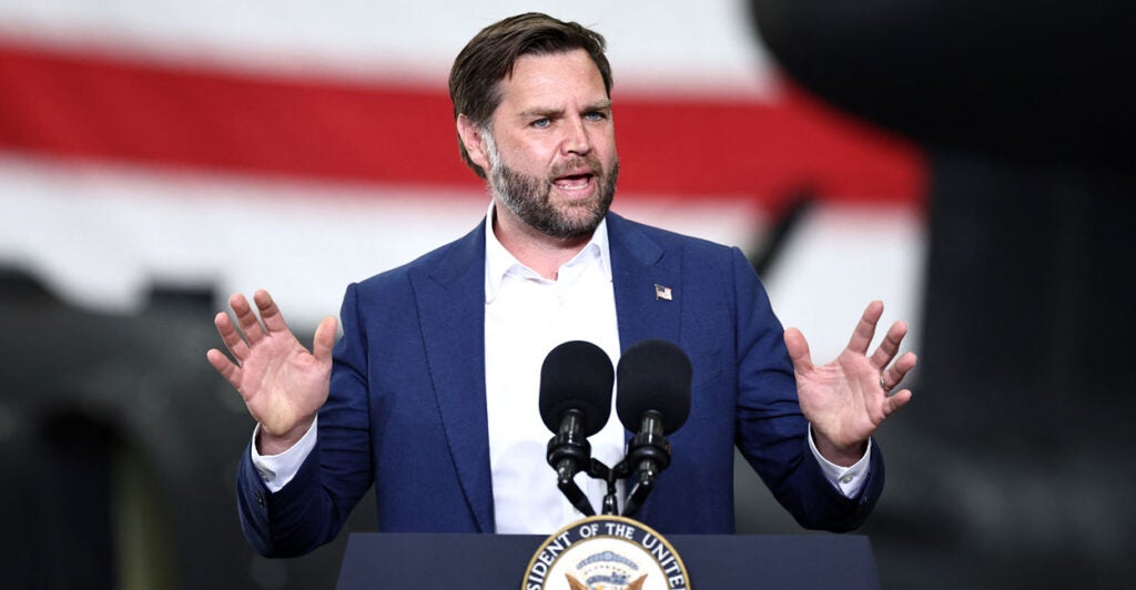 Vice President JD Vance at a podium, hands held shoulder high, with a large American flag behind him.