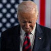 Joe Biden in a dark suit looking down at the floor with a large American flag behind him