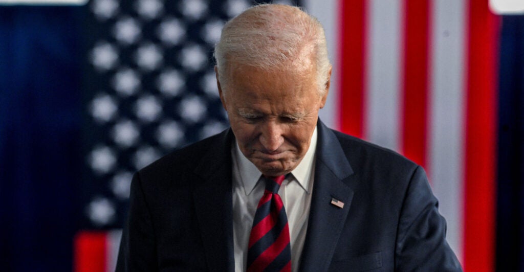 Joe Biden in a dark suit looking down at the floor with a large American flag behind him