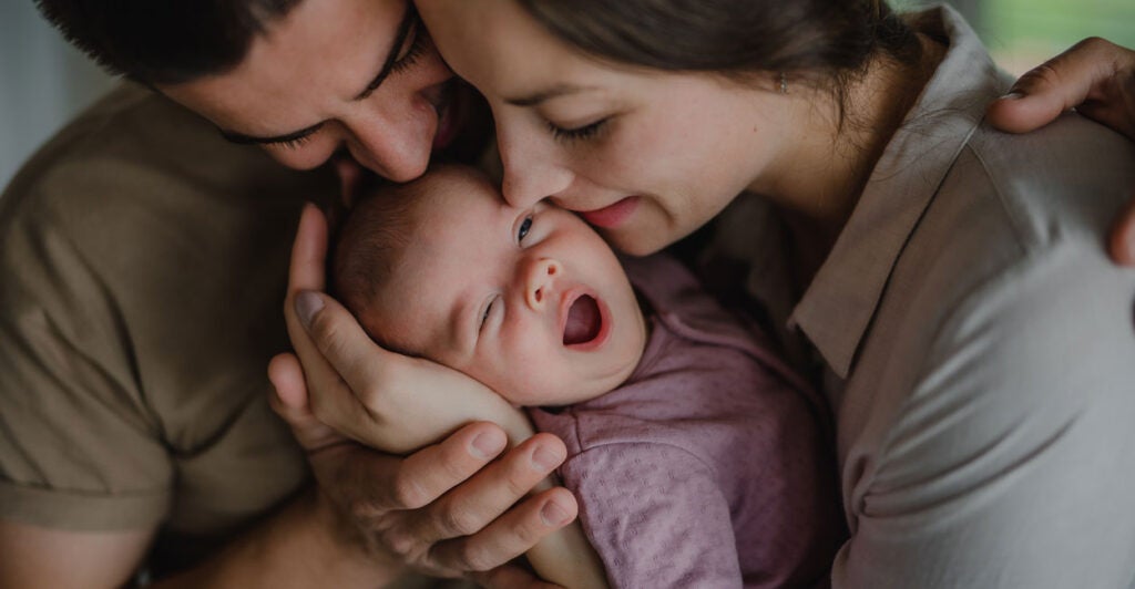 young parents holding and kissing their newborn baby indoors at home