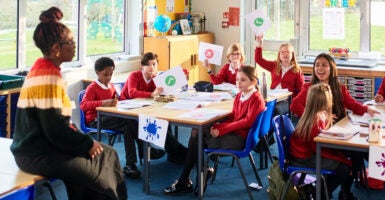 An elementary school class with children sitting around tables participating in classroom discussion wearing red sweaters