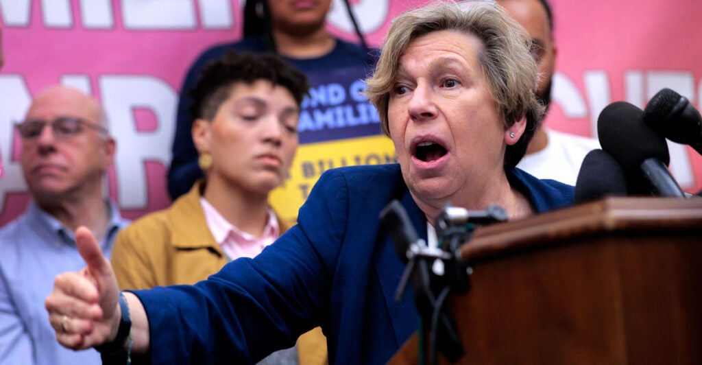 American Federation of Teachers President Randi Weingarten at a podium with multiple microphones speaking at a rally with rally goers behind her