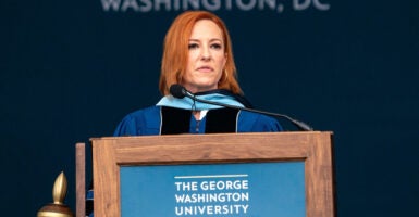 Jen Psaki wears a graduation gown as she stands behind a podium with a microphone as she looks out.