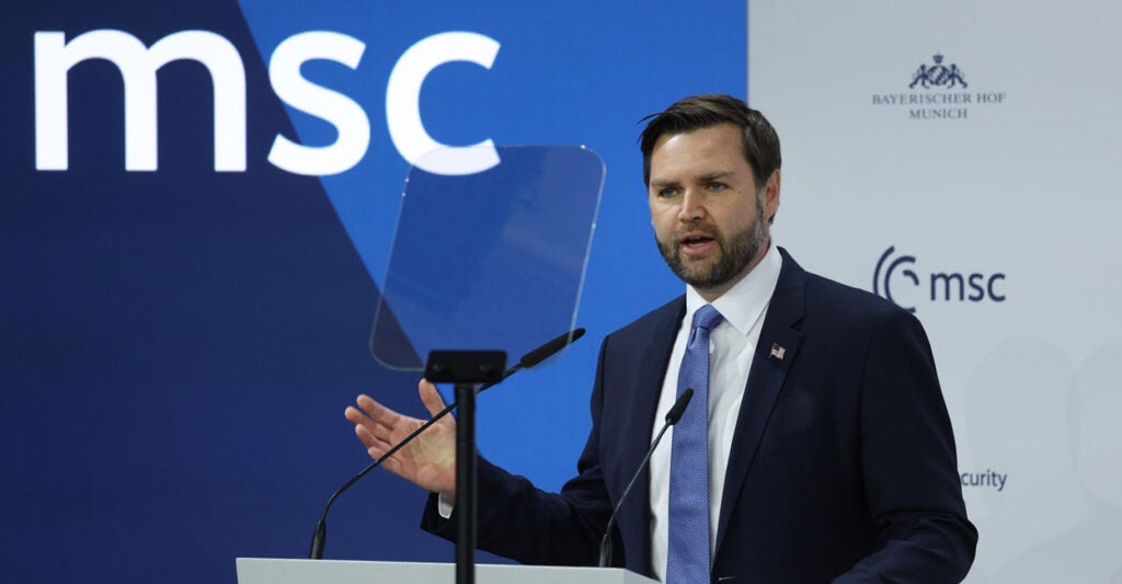 JD Vance sants before a podium and a microphone and speaks while holding out his right hand.