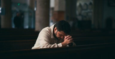A man kneels in a church with his hands clasped in front of him as he puts his head down in prayer.