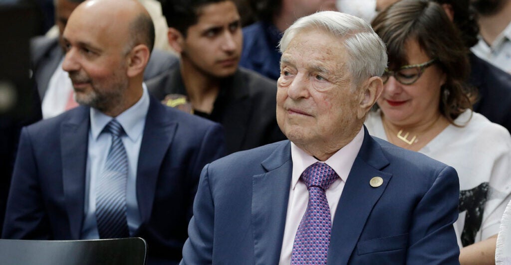 George Soros sits in a chair at an event and looks to ihs left.