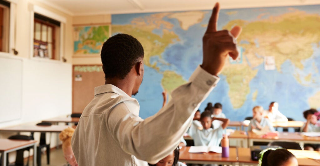 Black teacher in white shirt with back to the camera points to the sky while teaching young students.