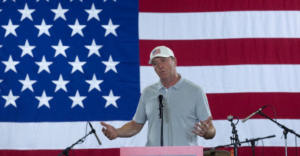 Sen. Eric Schmitt in light golf shirt and white hat with an American flag speaks from behind a podium. Behind him, a huge American flag.