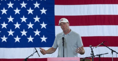 Sen. Eric Schmitt in light golf shirt and white hat with an American flag speaks from behind a podium. Behind him, a huge American flag.