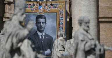 A portrait of late Pier Giorgio Frassati is displayed onto the facade of Saint Peter's Basilica. Statues in the foreground frame the portrait of the young man, arms folded, in a dark suit.