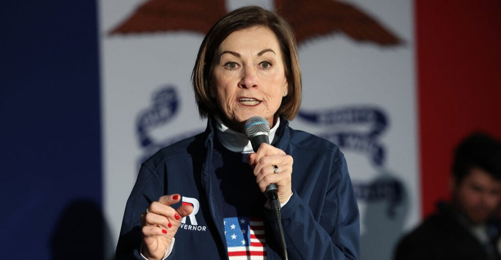 Iowa Gov. Kim Reynolds, in a blue sweater with American flag, speaks into a microphone.