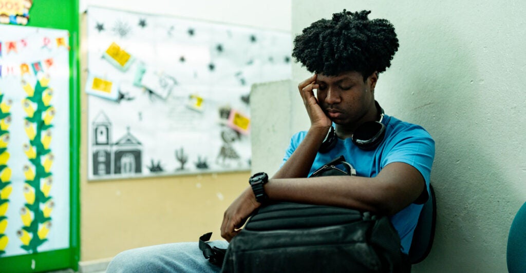 A black male teenage student sits against wall, clutching his backpack, head in hand, looking sad and worried.