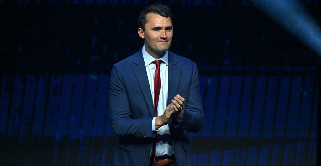 Charlie Kirk, in a blue suit against a blue background, clapping.
