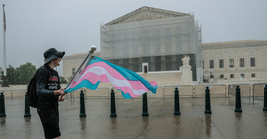 Protester carrying blue, pink, and white transgender flag walks in front of a scaffolded Supreme Court on a rainy day.