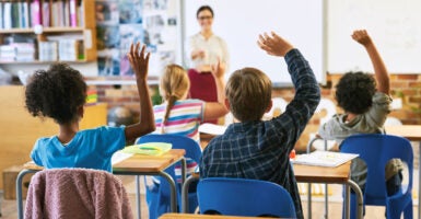 Grade-school kids raise their hands as teacher in white blouse and red dress speaks.