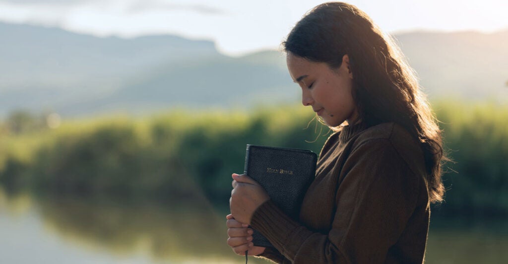 Young woman clutching a Bible prayers, in outdoor setting, with creek and mountains in background.
