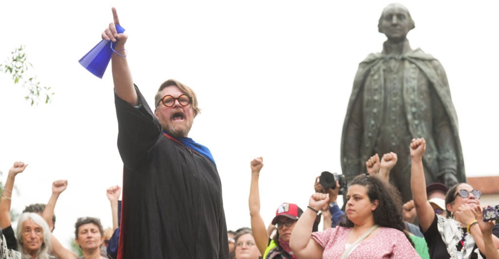 A bearded professor in black robe raises fist surrounded by students and faculty also with raised fist at May Day protest.