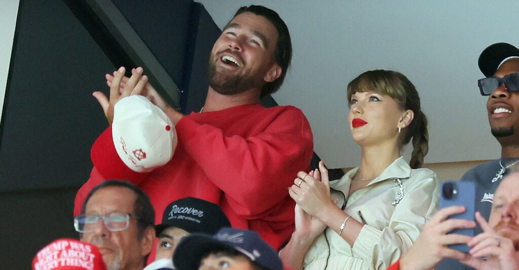 A jovial Travis Kelce in red shirt and smiling, clapping Taylor Swift enjoy a hockey game.